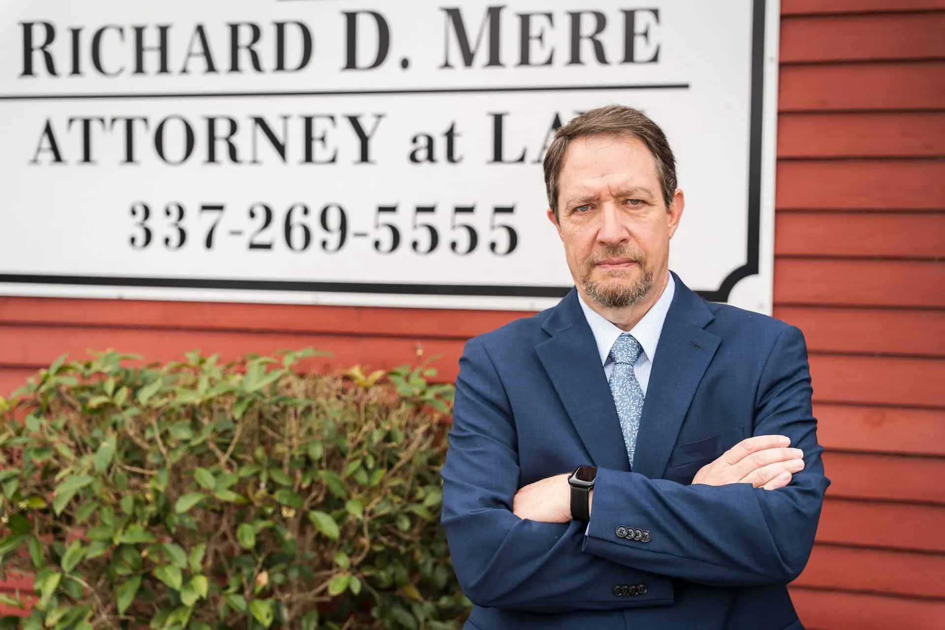 Richard D. Mere with his arms folded in front of the Richard D. Mere Attorney at Law sign outside his Lafayette office.